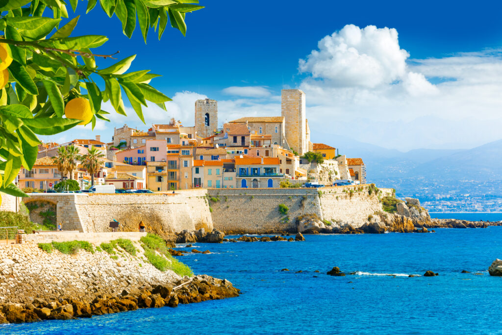 Pastellfarbene Häuser und Türme der Altstadt von Antibes thronen auf historischen Stadtmauern über dem türkisblauen Mittelmeer, vorn hängen Zitronenzweige.