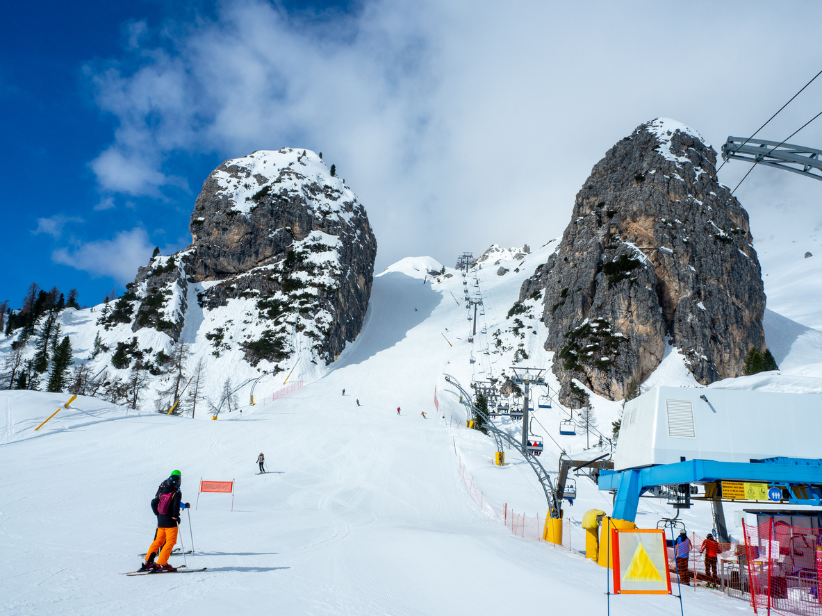 Sessellift und breite Piste mit Skifahrern vor markanten Felsformationen.
