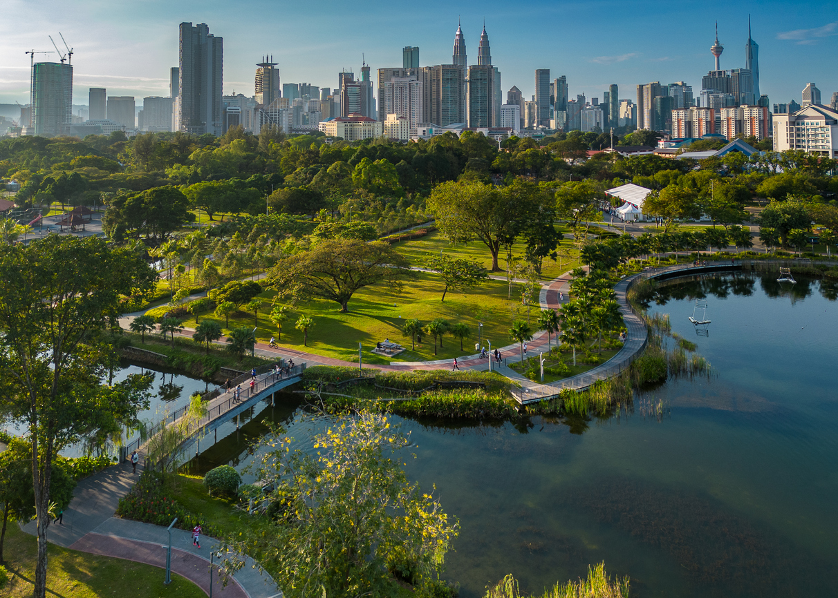 Grüner Stadtpark mit See, Stegen und Petronas Towers samt KL Tower im Hintergrund.
