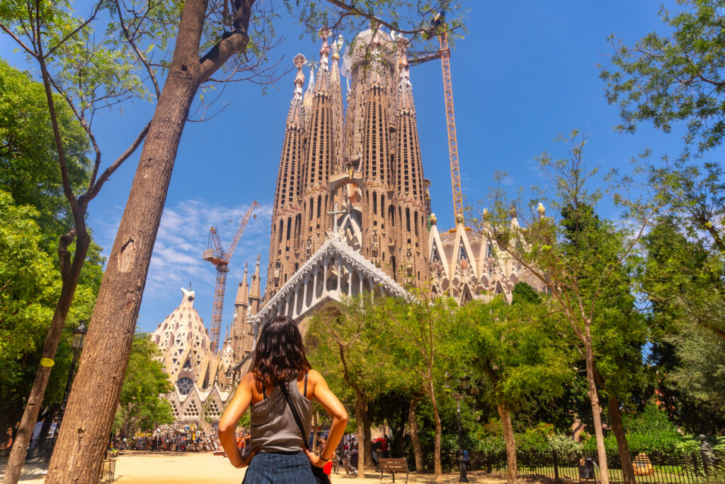 Eine Besucherin steht im Park vor der Sagrada Família in Barcelona, umgeben von Bäumen und Baukränen unter blauem Himmel.