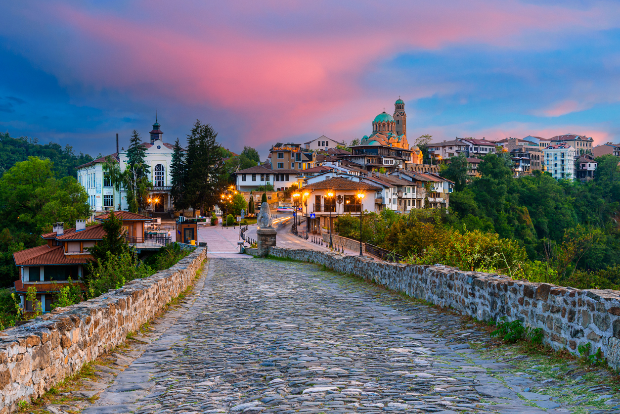 Kopfsteinpflaster-Brücke führt in die terrassenartige Altstadt von Veliko Tarnovo mit grün bekuppelter Kathedrale bei Abendhimmel.
