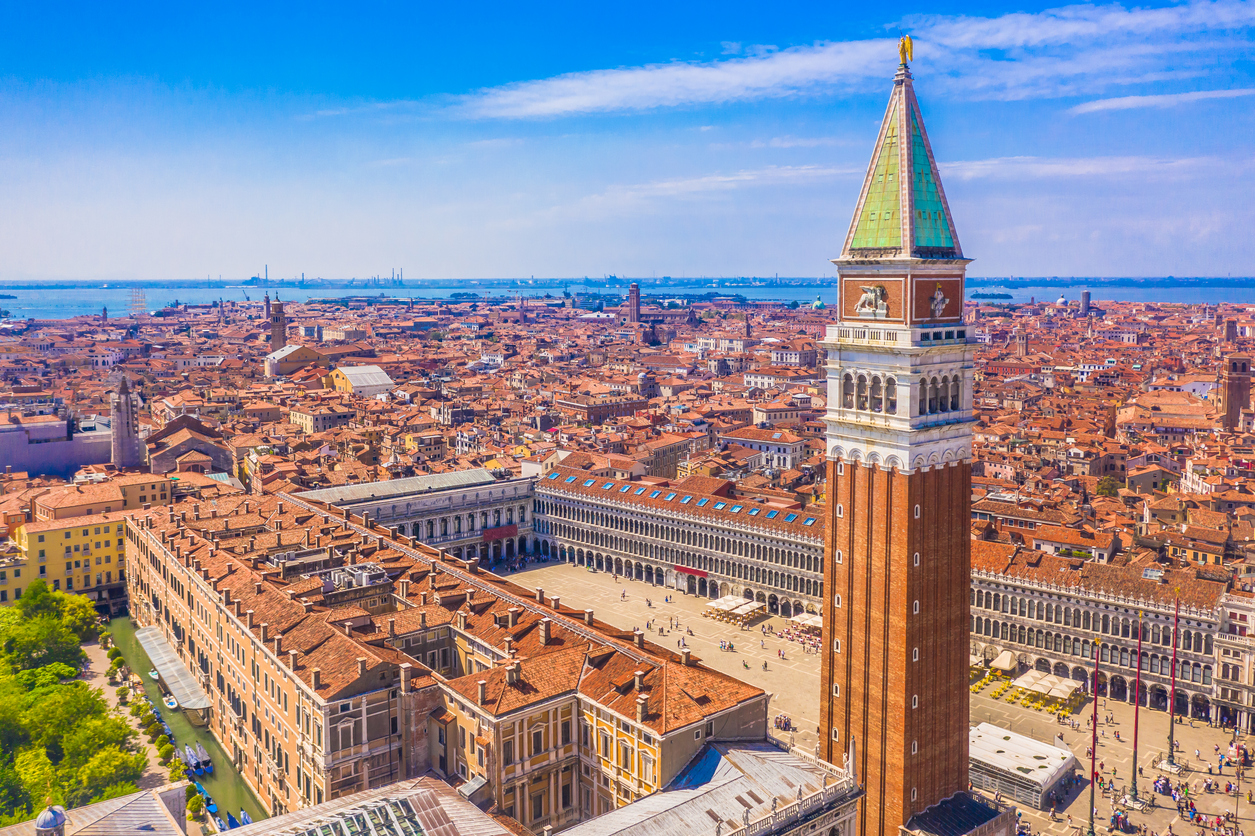 Luftaufnahme des Markusplatzes mit dem Campanile di San Marco, den Arkaden der Prokuratien und den roten Dächern bis zur Lagune unter blauem Himmel.