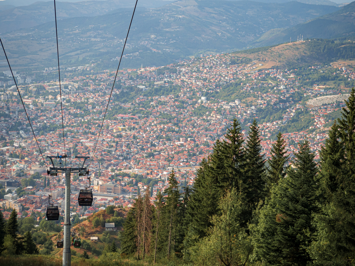 Gondeln der Trebević-Seilbahn gleiten über Nadelwald hinab auf Sarajewo mit dichtem Häusermeer im von Bergen umrahmten Tal.