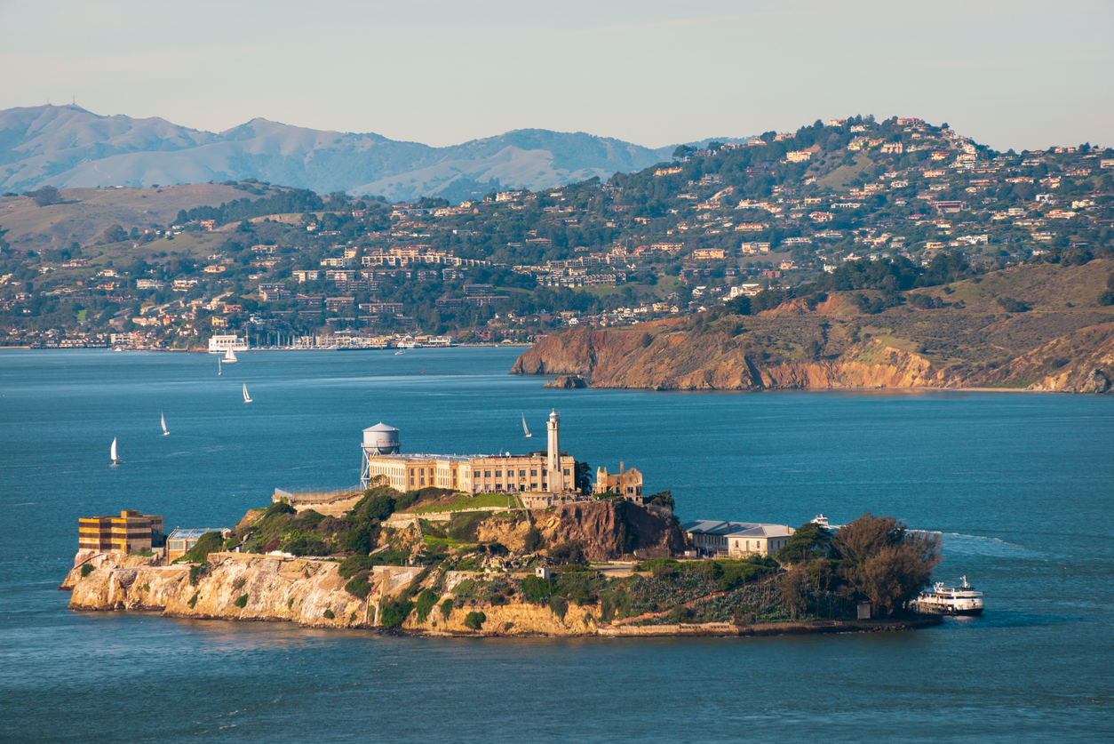 Blick auf Alcatraz Island mit Gefängniskomplex, Wasserturm und Leuchtturm in der Bucht von San Francisco, umgeben von blauem Wasser und Segelbooten.