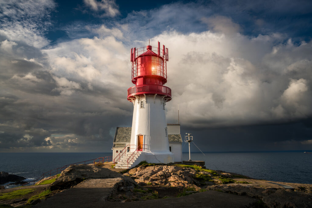Rot-weißer Leuchtturm auf Felsen am Meer unter dramatischen Wolken.