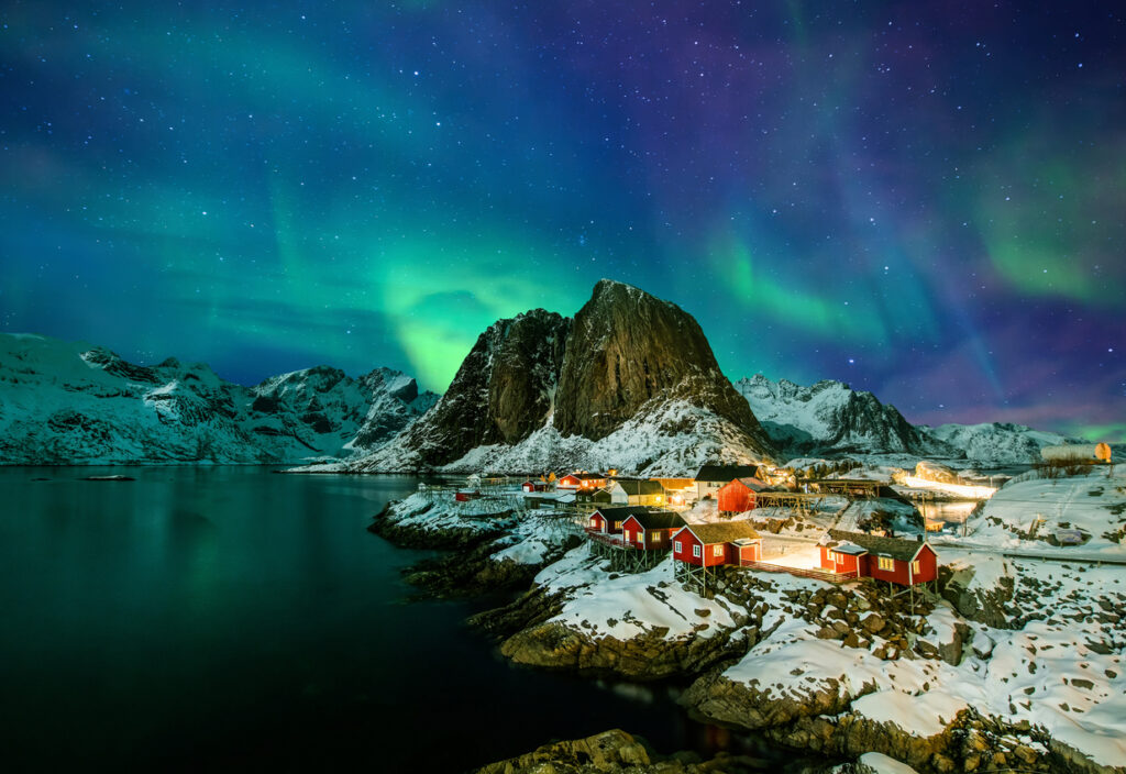 Grüne Aurora borealis über dem verschneiten Fischerdorf Hamnøy auf den Lofoten mit roten Rorbuer, steilen Bergen und Fjord.