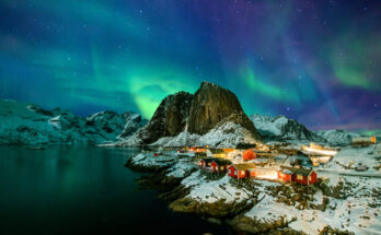 Grüne Aurora borealis über dem verschneiten Fischerdorf Hamnøy auf den Lofoten mit roten Rorbuer, steilen Bergen und Fjord.
