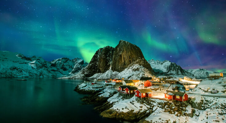 Grüne Aurora borealis über dem verschneiten Fischerdorf Hamnøy auf den Lofoten mit roten Rorbuer, steilen Bergen und Fjord.