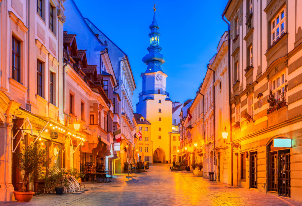 Blick durch eine leere Kopfsteinpflastergasse auf das beleuchtete Michaelertor mit Uhr, umgeben von historischen Häusern.