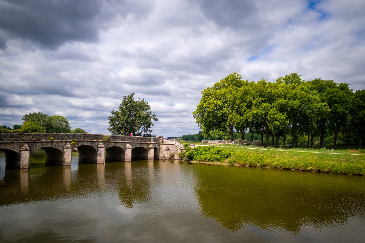 Steinerne Bogenbrücke über den Schlosskanal im Park von Chambord, mit Baumallee und Blickrichtung zum Château de Chambord.