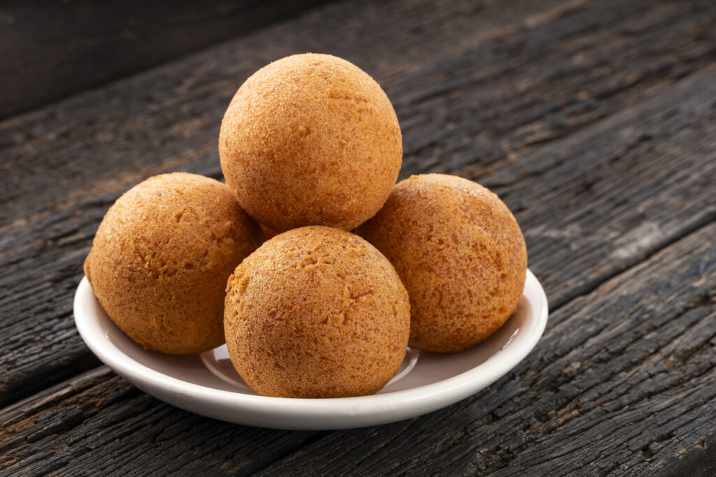 ünf goldbraune, runde Käse-Buñuelos liegen pyramidenförmig auf einem kleinen weißen Teller auf rustikalem Holz.
