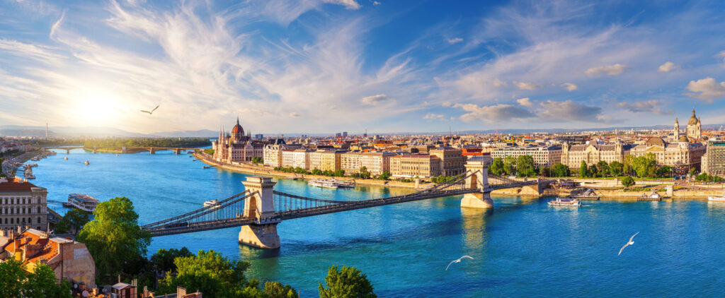Weitwinkelblick auf Budapest mit der Kettenbrücke über die Donau, dem ungarischen Parlament und historischen Fassaden bei blauem Himmel.
