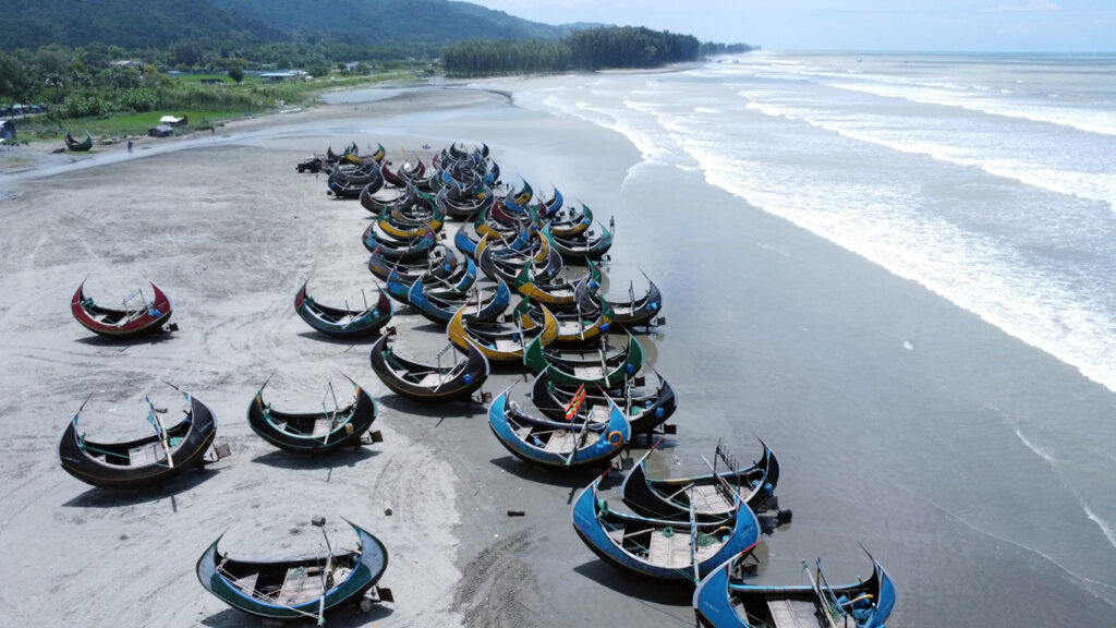 Dutzende sichelförmige Holzboote liegen bei Ebbe auf dem Strand von Cox’s Bazar mit Brandung des Golfs von Bengalen und grünen Hügeln im Hintergrund.