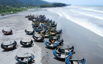 Dutzende sichelförmige Holzboote liegen bei Ebbe auf dem Strand von Cox’s Bazar mit Brandung des Golfs von Bengalen und grünen Hügeln im Hintergrund.