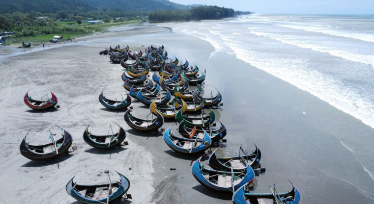Dutzende sichelförmige Holzboote liegen bei Ebbe auf dem Strand von Cox’s Bazar mit Brandung des Golfs von Bengalen und grünen Hügeln im Hintergrund.