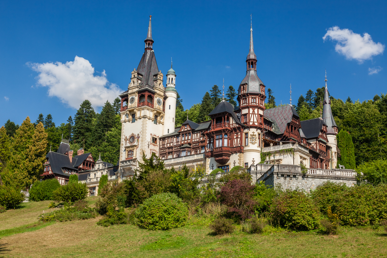 Schloss Peleș mit hohen Türmen, Fachwerk und Schieferdächern vor bewaldeten Karpaten und blauem Himmel.