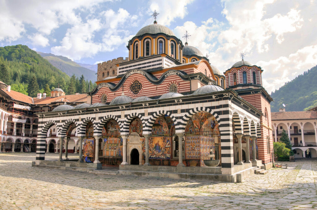 Hauptkirche des Rila-Klosters mit schwarz-weißen Arkaden und farbigen Fresken vor bewaldeten Berghängen.