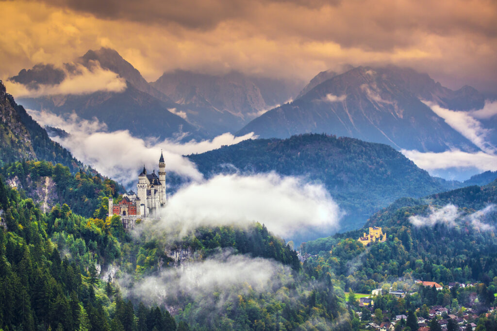 Panorama mit Schloss Neuschwanstein auf bewaldetem Felsen, umgeben von Wolken und den Ammergauer Alpen.