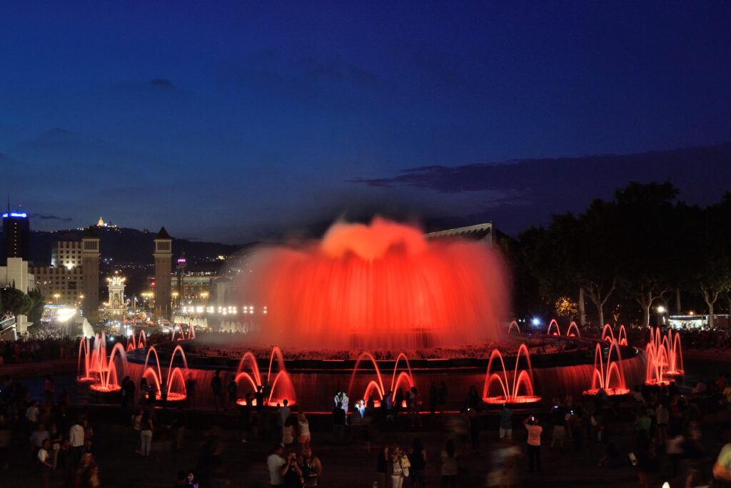 Nachtaufnahme des Font Màgica de Montjuïc mit rot illuminierten Wasserfontänen und Zuschauerreihen, dahinter die Venezianischen Türme am Plaça d’Espanya.