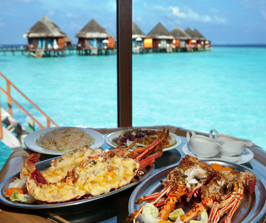 Gegrillter Hummer mit Reis und Salat vor türkisblauer Lagune und Wasserbungalows.