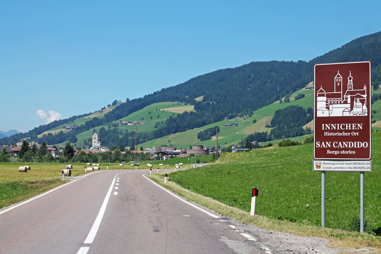 Straßenschild „Innichen / San Candido – Historischer Ort“ vor grünen Hängen und Dorf.