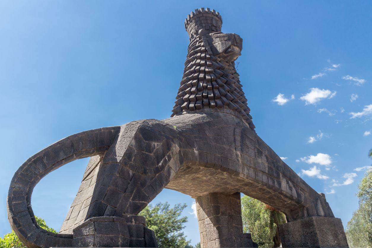 Monumentale, dunkelsteinige Löwenstatue (Löwe von Juda) nahe dem Nationaltheater in Addis Abeba; stilisierter Löwe mit erhobenem Kopf, schuppenartiger Mähne und ringförmig geschwungenem Schwanz, vor blauem Himmel und Bäumen.