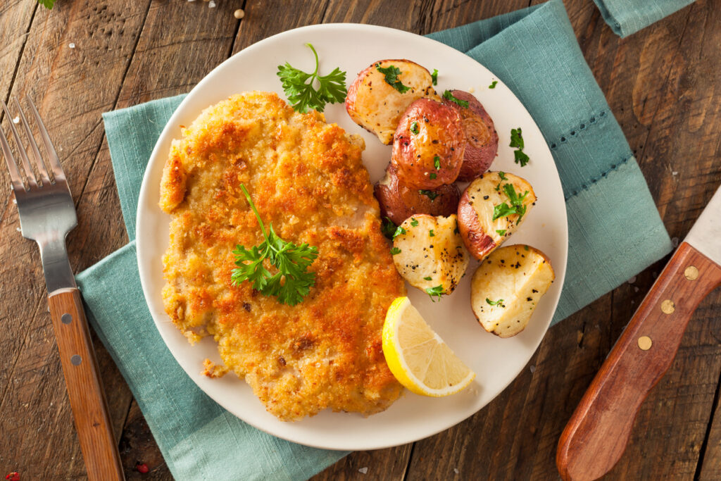 Goldbraunes paniertes Schnitzel mit Zitronenspalte und Bratkartoffeln auf weißem Teller.