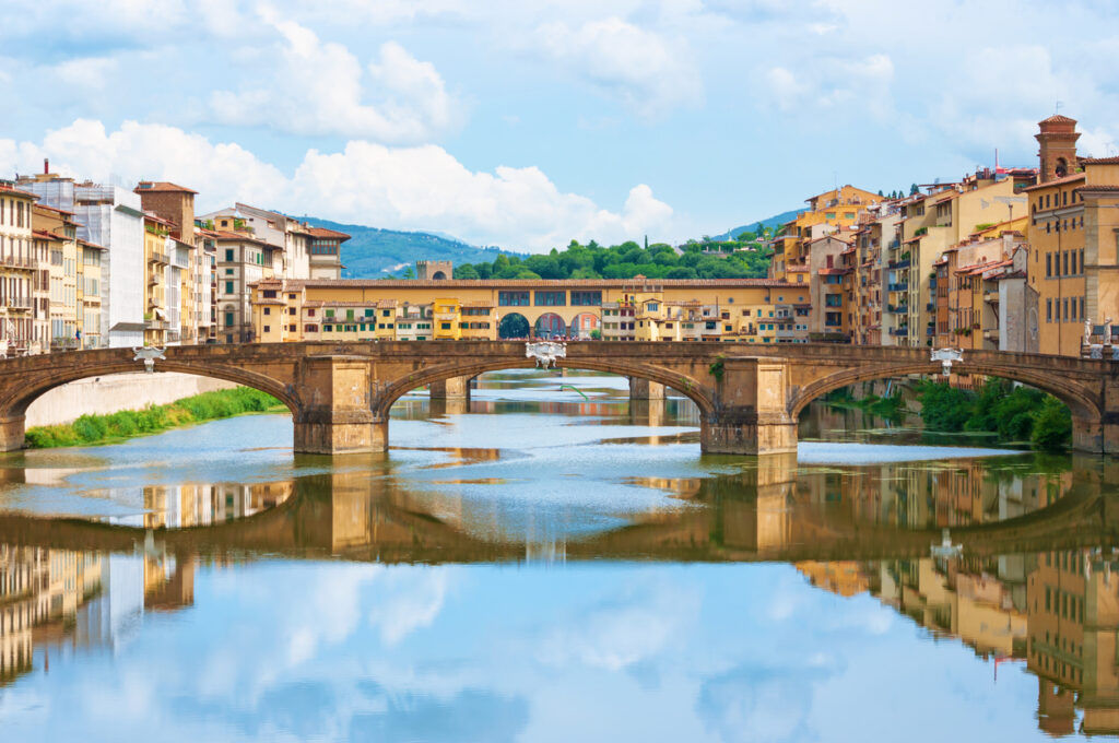 Blick auf die Ponte Vecchio in Florenz mit beidseitigen Häuserzeilen und Spiegelung im Arno.