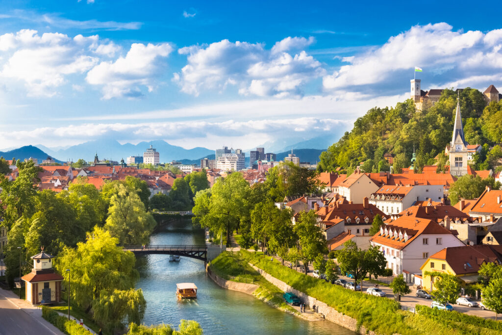 Blick über die Ljubljanica mit Brücken, roten Dächern und Burghügel in sommerlichem Licht.