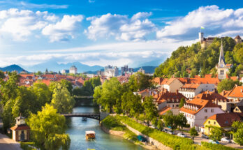 Blick über die Ljubljanica mit Brücken, roten Dächern und Burghügel in sommerlichem Licht.