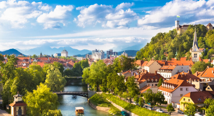 Blick über die Ljubljanica mit Brücken, roten Dächern und Burghügel in sommerlichem Licht.