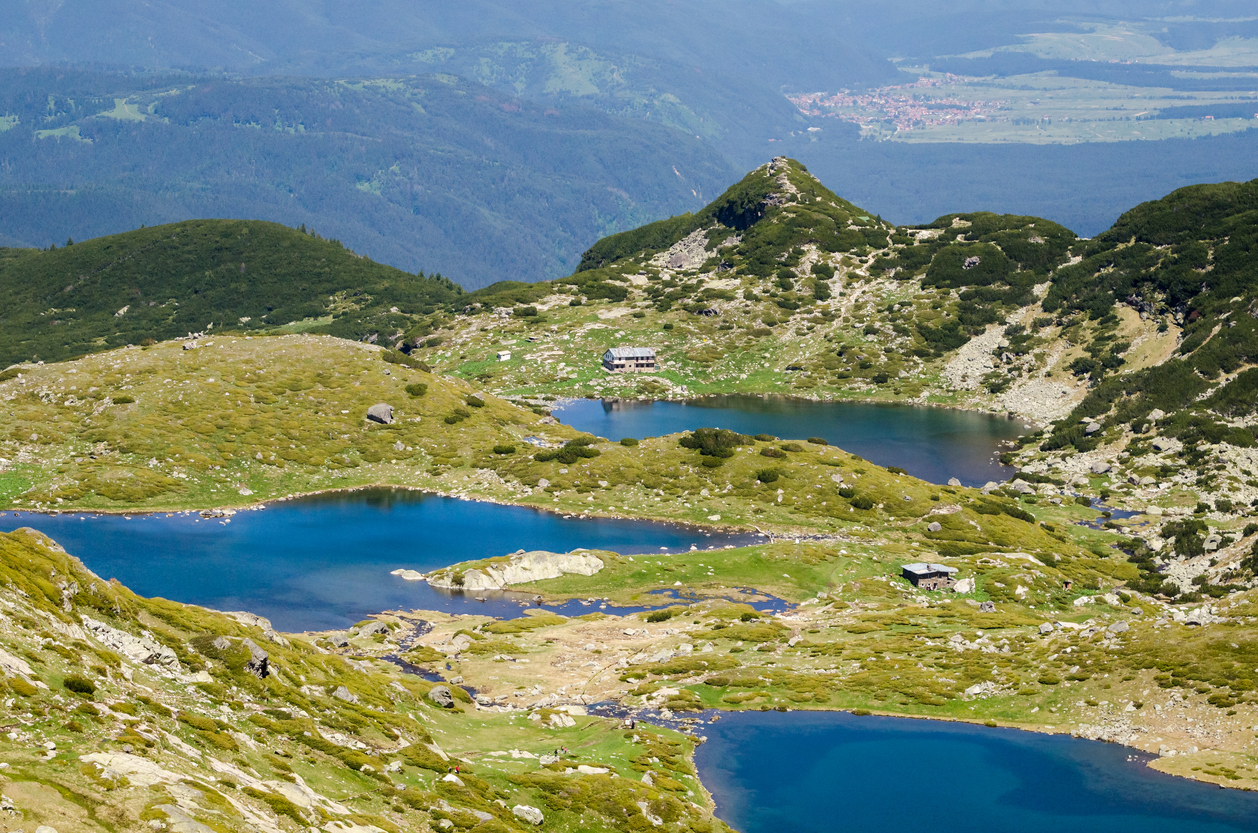 Türkisblau schimmernde Gletscherseen liegen zwischen grünen Hängen und zwei Berghütten in hochalpiner Landschaft.