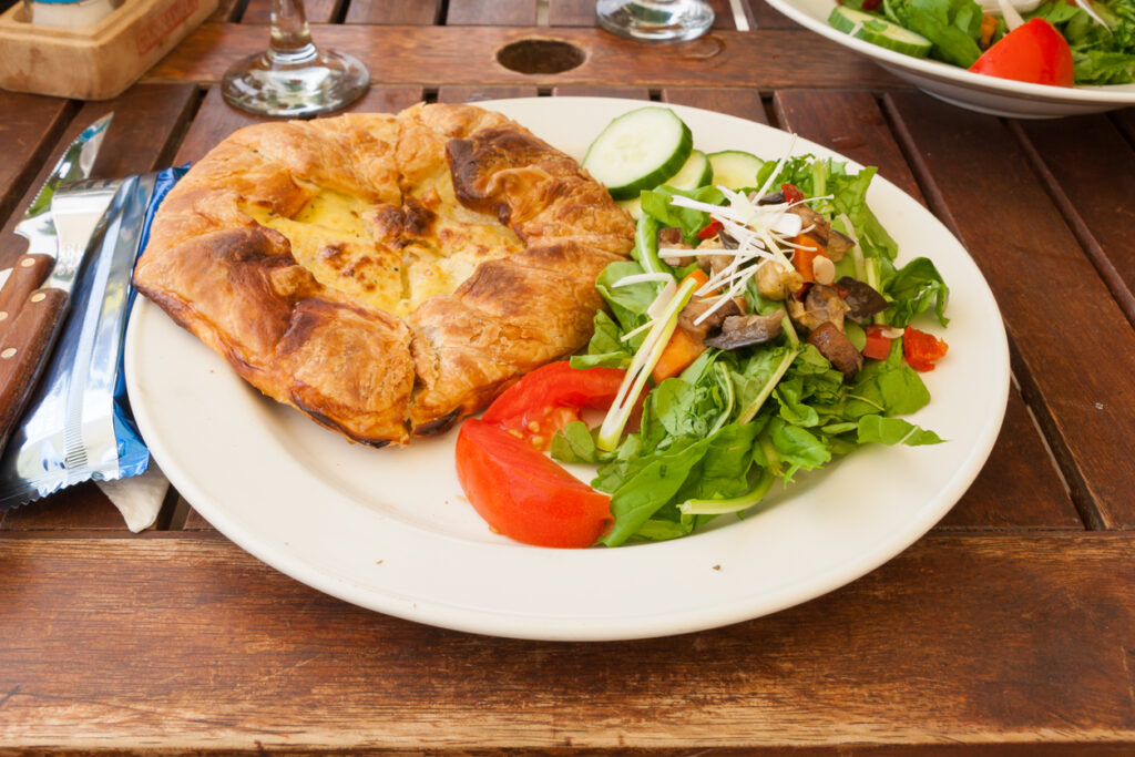 Teller mit goldbrauner Blätterteig-Pastete und gemischtem Salat aus Rucola, Gurke und Tomate auf Holztisch.