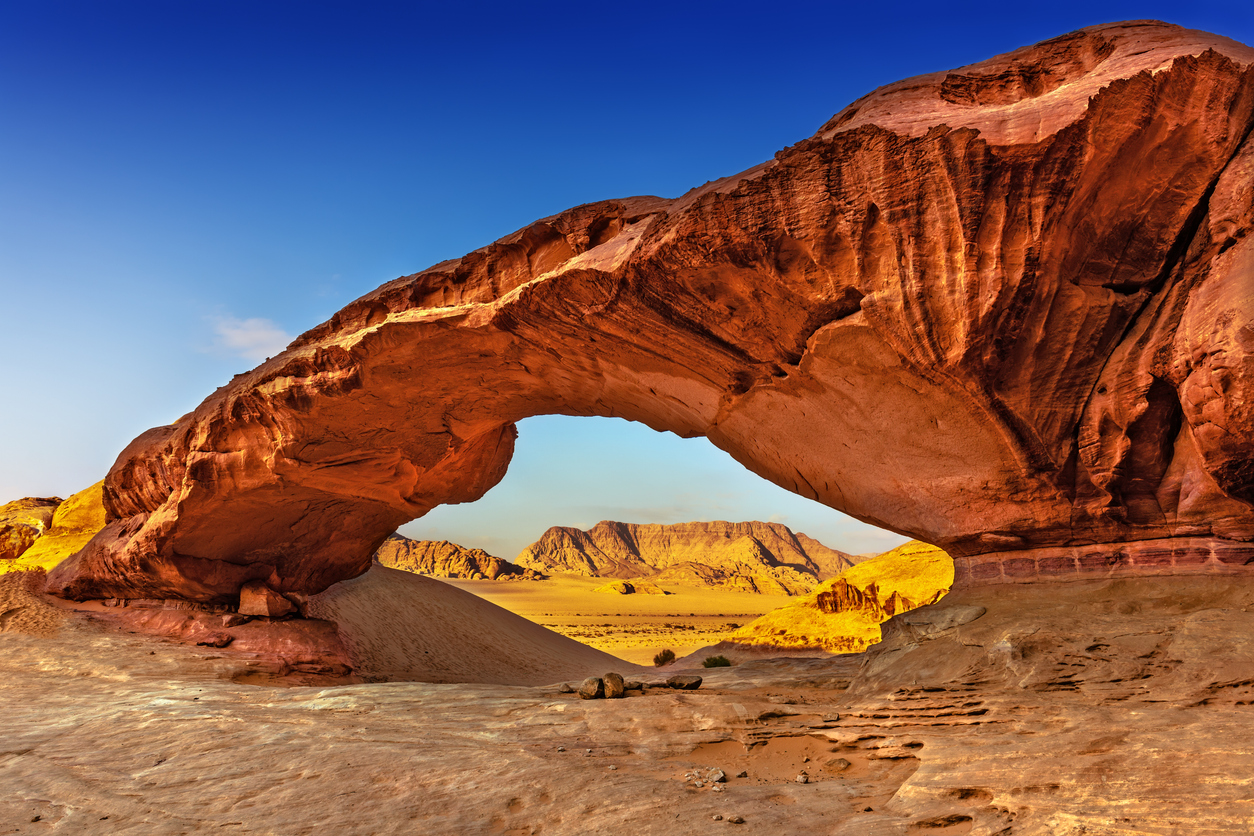 Roter Sandstein-Felsbogen im Wadi Rum mit Bergen und goldenem Wüstenpanorama dahinter.