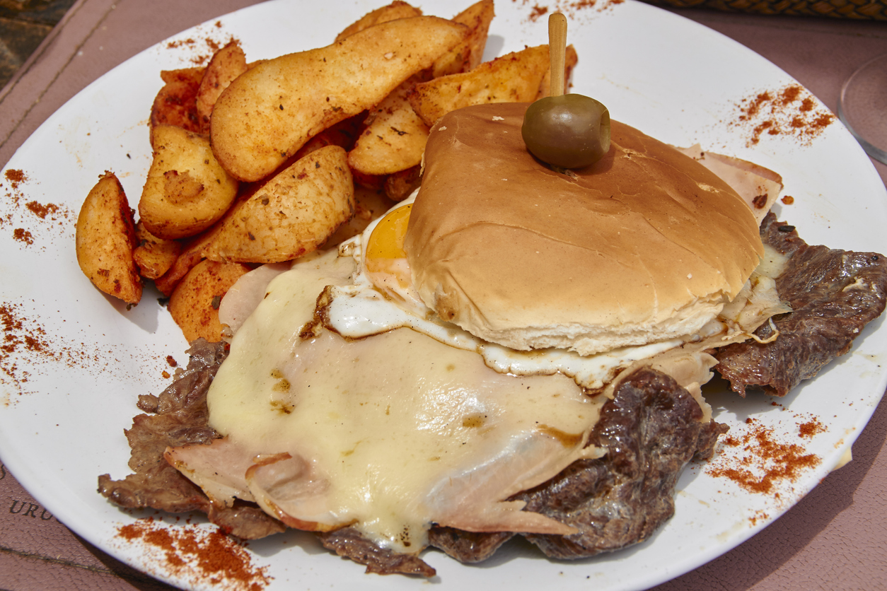 Chivito-Sandwich mit Steak, Schinken, Käse und Spiegelei, serviert mit gewürzten Kartoffelspalten auf weißem Teller.
