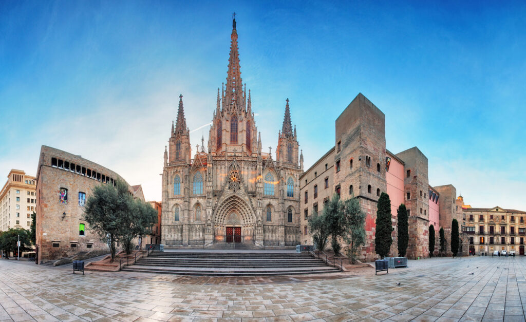 Weitwinkelansicht der gotischen Kathedrale von Barcelona mit leerem Platz, Treppen und mittelalterlichen Mauern unter blauem Himmel.