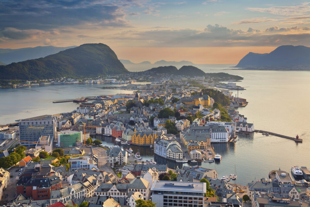 Luftaufnahme von Ålesunds Altstadt mit farbigen Jugendstilfassaden am Hafen, umgeben von Inseln, Bergen und Meer im warmen Abendlicht.