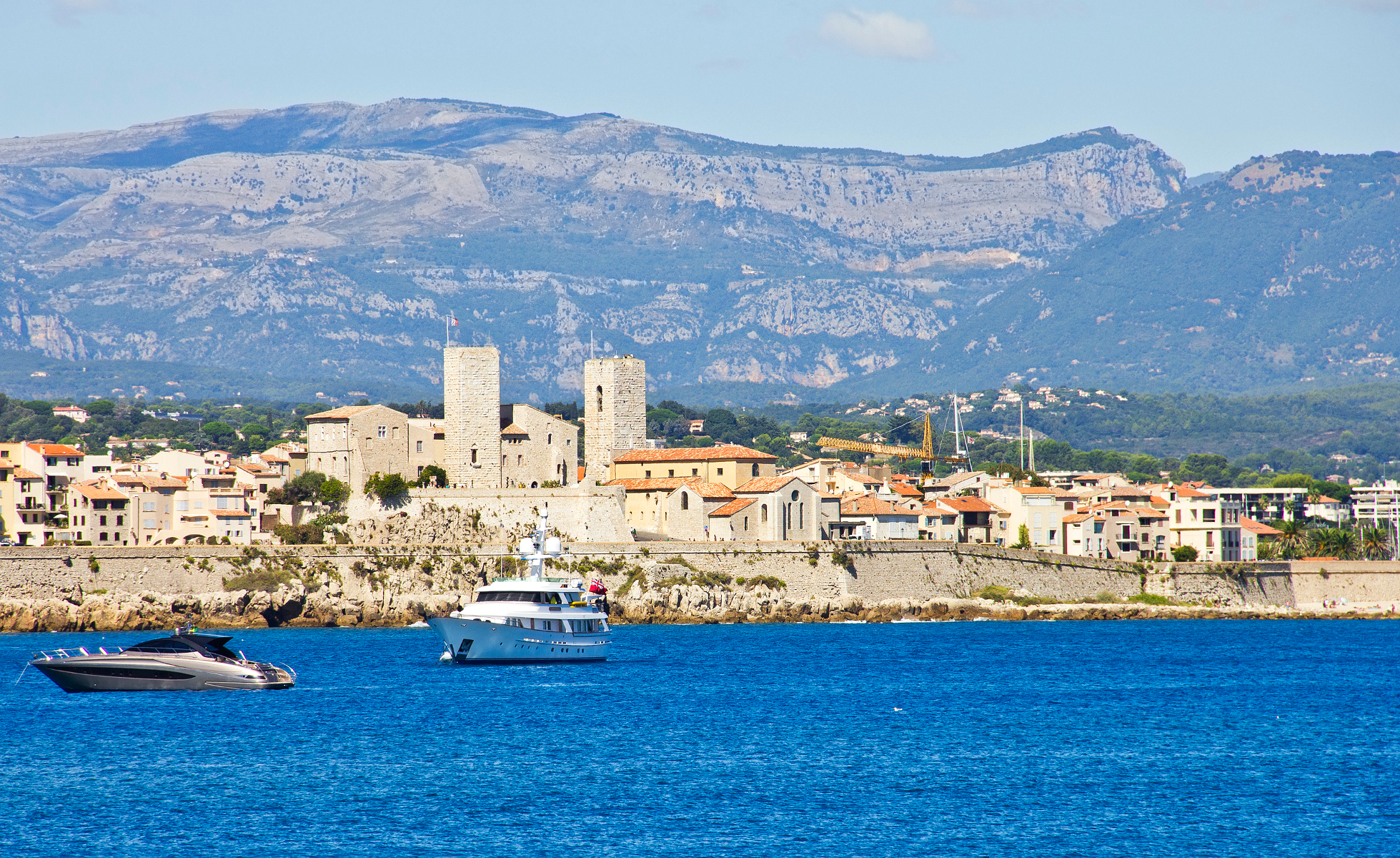Blick über das Meer auf Antibes mit steinernen Stadtmauern und zwei markanten Türmen der Altstadt, davor kreuzen Yachten im türkisblauen Wasser, im Hintergrund erheben sich die Seealpen.