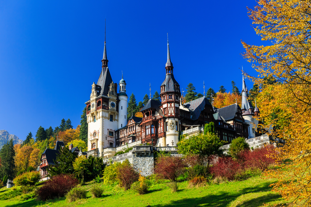 Blick auf Schloss Peleș mit hohen Türmen und Fachwerk, umgeben von buntem Herbstwald und blauem Himmel.
