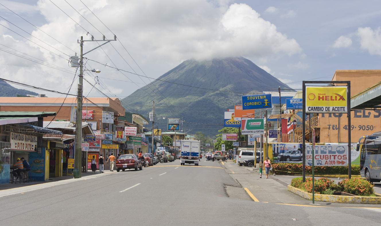 Hauptstraße mit bunten Läden und Schildern; dahinter der Vulkan Arenal mit Wolkenkappe.