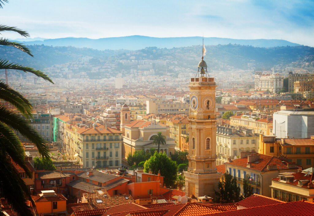 Panoramablick über die Altstadt von Nizza mit ockerfarbenen Fassaden und Terrakottadächern, einem markanten Uhrturm, Palmen im Vordergrund und den dunstigen Hügeln der Seealpen im Hintergrund.