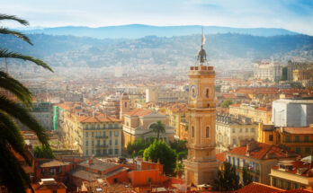 Panoramablick über die Altstadt von Nizza mit ockerfarbenen Fassaden und Terrakottadächern, einem markanten Uhrturm, Palmen im Vordergrund und den dunstigen Hügeln der Seealpen im Hintergrund.