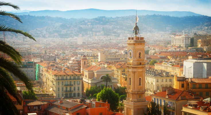 Panoramablick über die Altstadt von Nizza mit ockerfarbenen Fassaden und Terrakottadächern, einem markanten Uhrturm, Palmen im Vordergrund und den dunstigen Hügeln der Seealpen im Hintergrund.