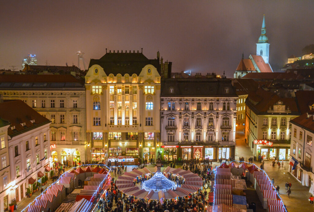 Luftaufnahme eines beleuchteten Weihnachtsmarkts mit rot-weißen Ständen auf einem Platz in Bratislava, umgeben von historischen Fassaden.