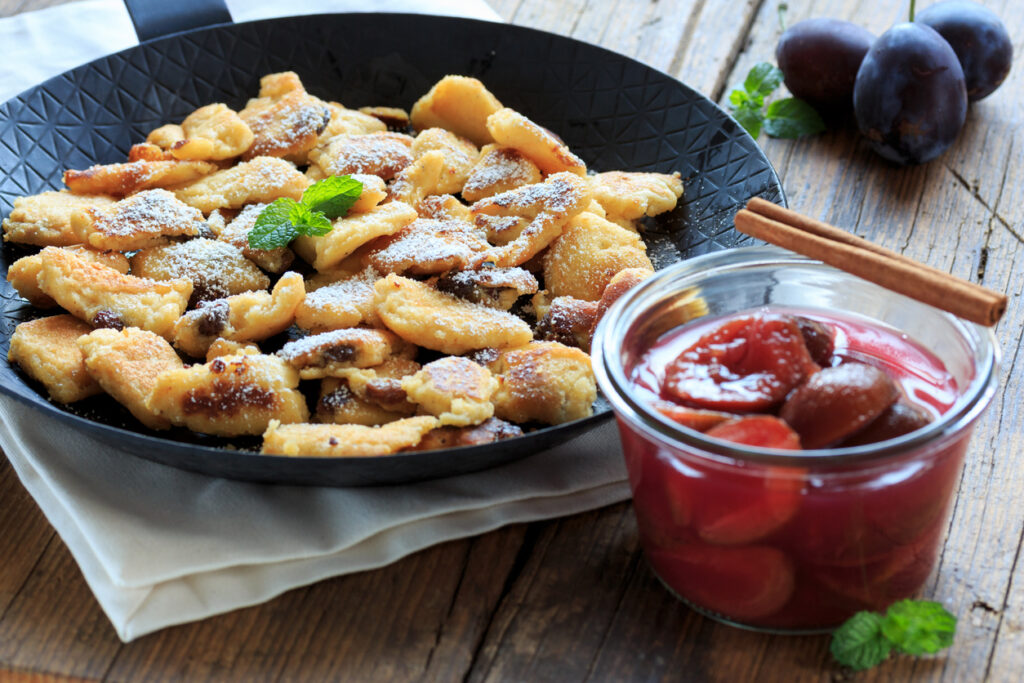 Goldbrauner Kaiserschmarrn in einer Pfanne mit Puderzucker, daneben ein Glas Zwetschgenröster mit Zimtstange auf Holztisch.