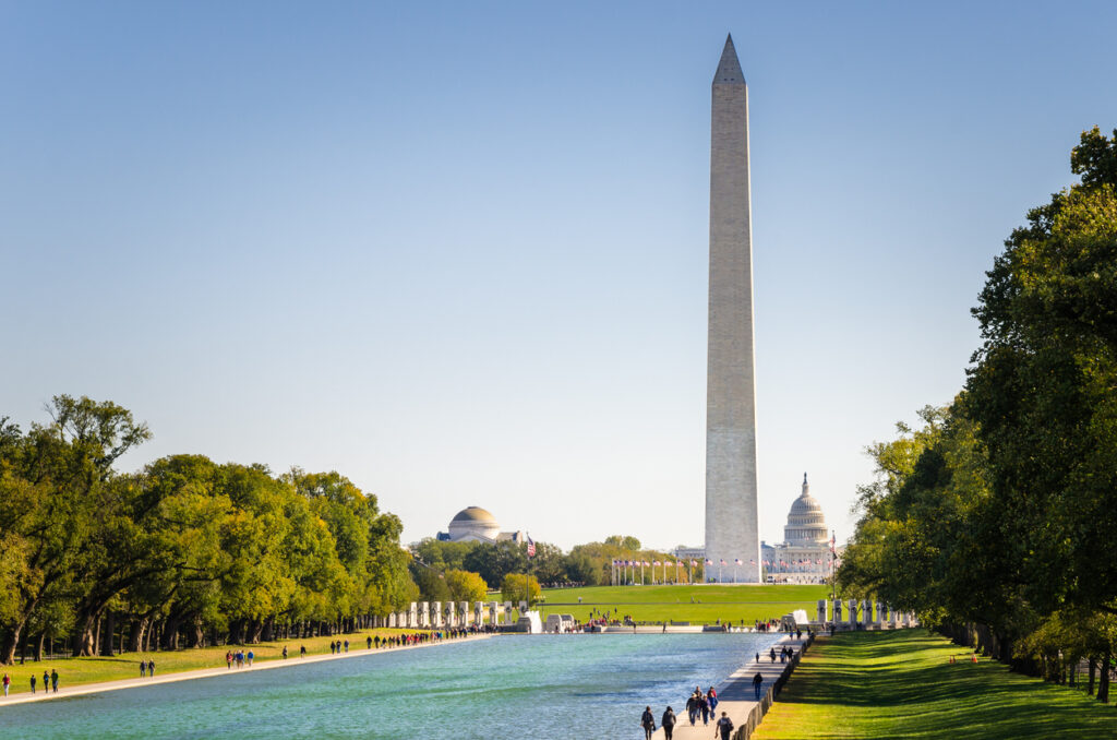 Das Washington Monument erhebt sich über dem Reflecting Pool, flankiert von Bäumen, mit dem Kapitol dahinter.