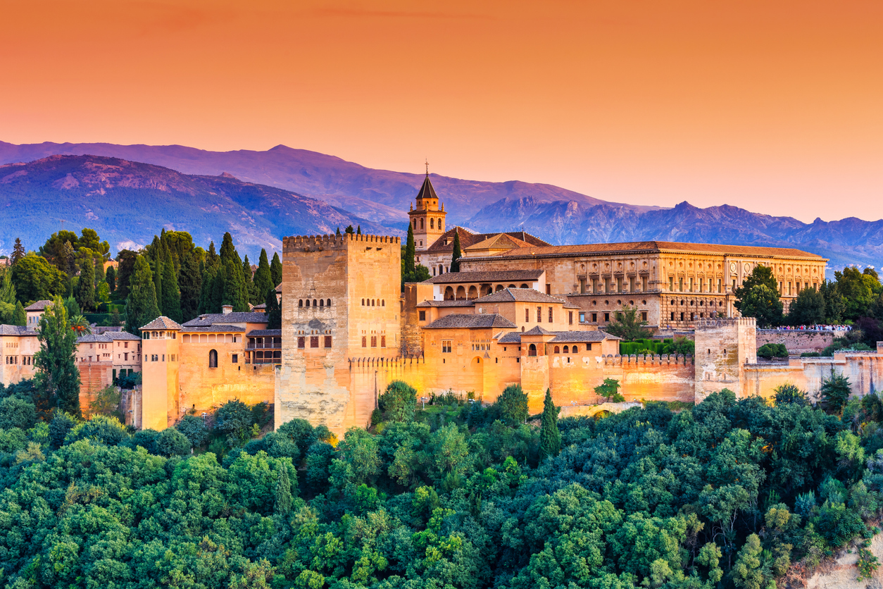 Panorama der Alhambra in Granada mit Palästen und Zinnen über grünen Zypressen vor orangefarbenem Himmel.