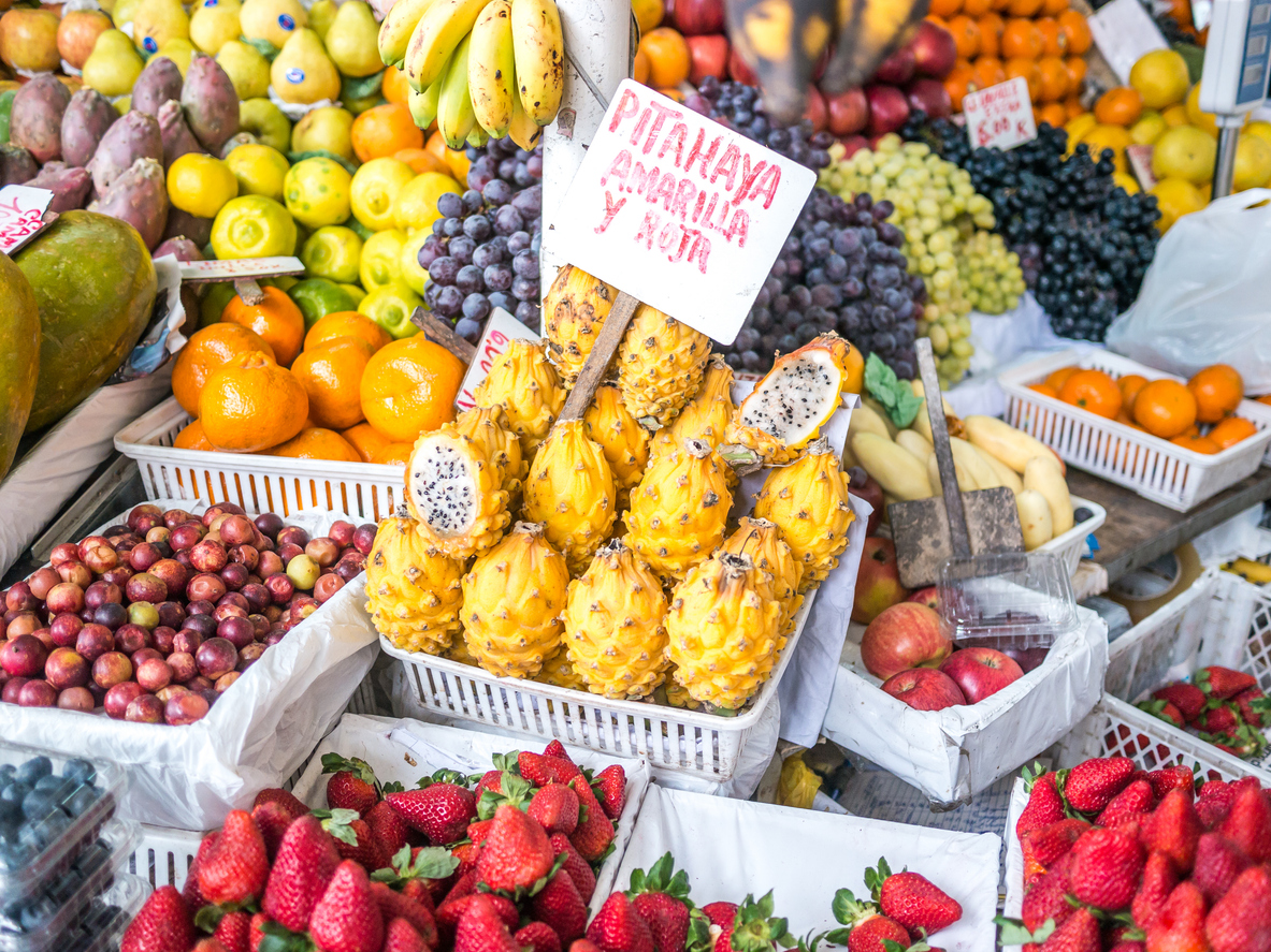 Bunte Obststände mit gelber Pitahaya, Erdbeeren, Trauben, Zitrusfrüchten und handgeschriebenen Schildern auf einem Markt in Peru.