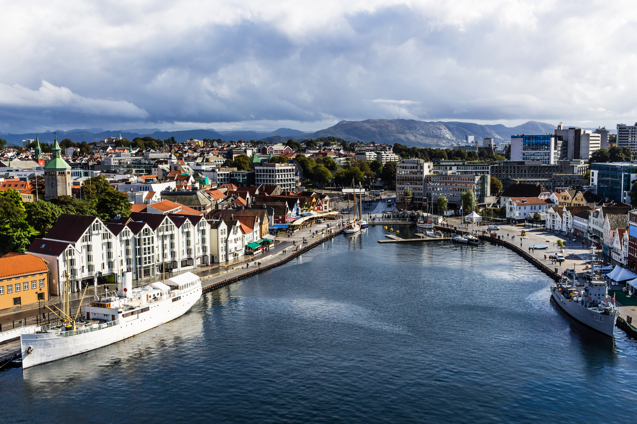 Panorama des Hafens von Stavanger mit Schiffen, Uferpromenade, weißen Holzfassaden und Bergen im Hintergrund unter wechselhaftem Himmel.