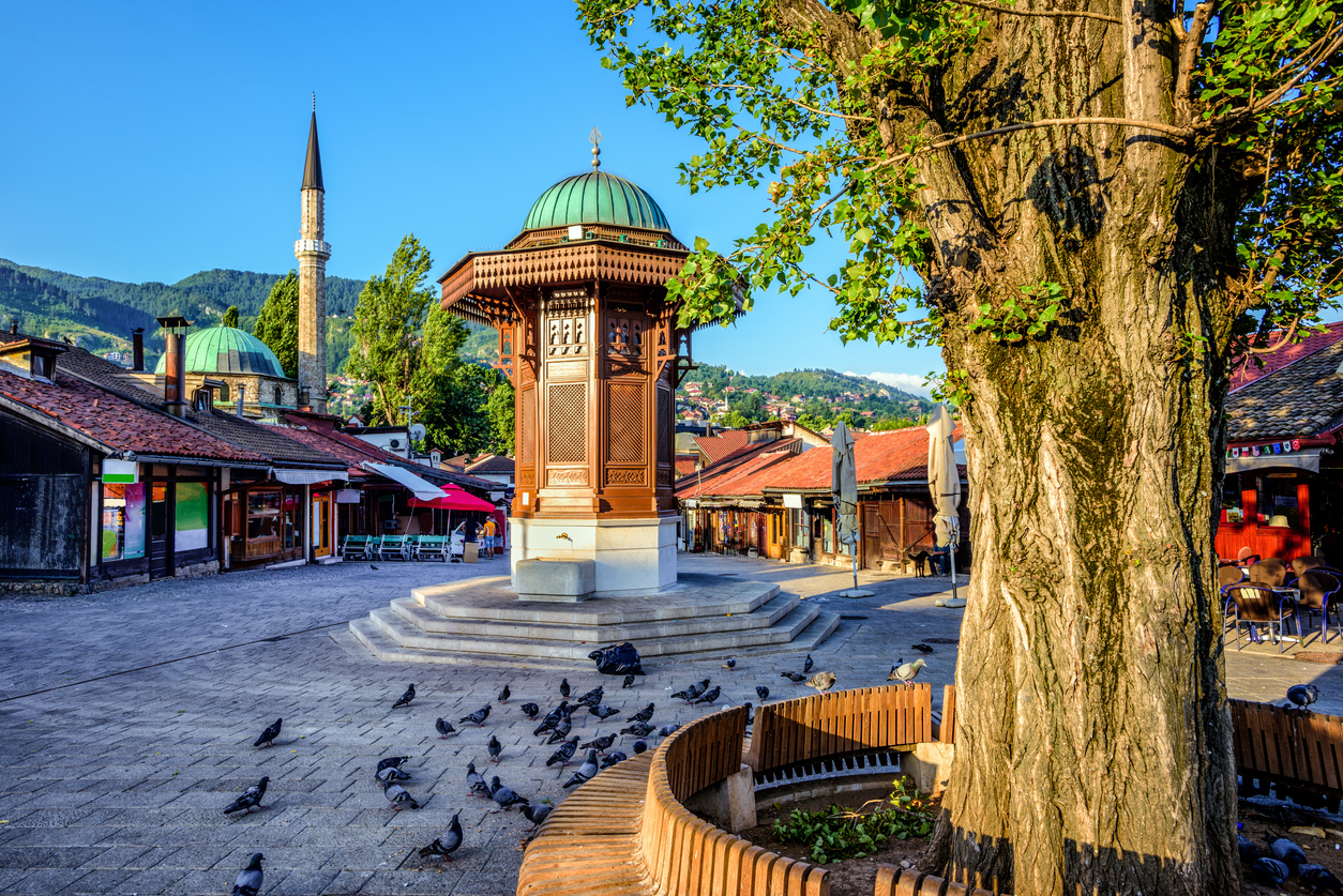 Sebilj-Holzbrunnen in Sarajewos Baščaršija mit Minarett, Altstadthäusern, Bergen und vielen Tauben unter blauem Himmel.